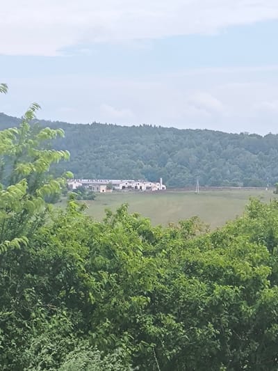 A long, white building complex with a red roof is situated in a grassy field in Holčíkovce, Slovakia, framed by dense green foliage in the foreground and a treed hill in the background. The building appears to be a farm or industrial facility, with multiple sections and a tall, slender structure resembling a silo or smokestack on its right side. A power line pole stands in the field in front of the building. The sky above is cloudy and a pale blue.
