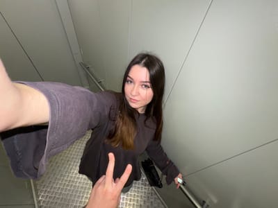 A young woman with long, dark brown hair and blue eyes is taking a selfie on a metallic staircase. She is wearing a dark gray long-sleeved shirt. Her left arm is extended, holding the camera, and her right hand is near her chest, forming a peace sign. A black quilted handbag with a chain strap is visible on her right side. The staircase has a textured metal floor and a silver railing. The walls are a light gray color, with visible seams. The scene appears to be indoors, possibly in a modern building or public transport. The specific location in Shirkovo, Russia, is not identifiable from the visual information in the image.