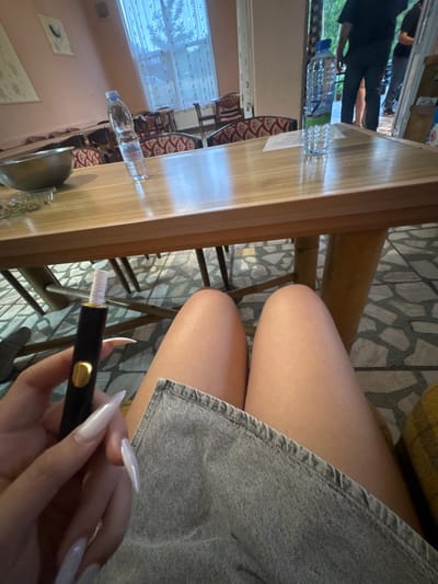 A person is sitting at a wooden table, possibly in a restaurant or home in Ustrem, Bulgaria. The location is suggested by the presence of a bottle of "Predela" brand water, which is a Bulgarian brand. The person is wearing a light grey denim skirt or shorts. In their hand, they are holding a black and gold object, possibly a vape pen or similar device. The person has long, pointed, white painted nails. On the table, there is a metal bowl and what appears to be some greenery, perhaps a garnish. In the background, a window with white curtains is visible, offering a glimpse of the outdoors. The floor is tiled with a geometric pattern.