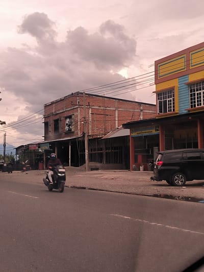 The image depicts a street scene in Ujong Duabelas, Indonesia, under an overcast sky with warm, reddish-pink hues, suggesting either sunrise or sunset. In the foreground, an asphalt road stretches, with a person riding a black scooter away from the viewer. The rider is wearing a helmet or mask.
Along the right side of the road, various buildings line the street. On the far right is a brightly painted two-story structure with a yellow upper facade and a light blue lower section, featuring white-framed windows, one of which is open. Next to it, a black SUV is parked. Above a shopfront adjacent to the SUV, a sign reads "Agar Neaga Jamurum" (or similar, with "Agar Neaga" being clearer), with smaller, illegible text below.
Towards the center-left, a multi-story building appears to be either under construction or in a state of disrepair, characterized by exposed red brick, rebar, and broken windows. Overhead utility lines crisscross the sky in front of these buildings. Further to the left, a sign reads "JUAL BAN BEKAS" (meaning "SELL USED TIRES"), situated near a stack of tires. In the distant background on the left, mountains are visible beneath the cloud-covered sky. The ground on the roadside is unpaved, showing gravel and dirt. The overall scene captures a slice of daily life in a bustling, somewhat rustic commercial area.