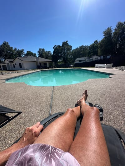 The image depicts a person relaxing by a pool on a sunny day. The perspective is from the person's point of view, looking down at their legs and the pool. They are seated on a black lounge chair, wearing patterned swim trunks and flip-flops. Their legs are tanned and appear to have been in the sun.

The scene is a backyard with a swimming pool, surrounded by a pebbled concrete deck. A large green tree with dark foliage is visible in the background, along with a white modern house. The sky is a clear, bright blue, indicating fair weather and likely daytime, judging by the sunlight and shadows. A faint lens flare is visible in the upper left corner of the image, a common characteristic of sunny outdoor photography. In the far background, a white RV or trailer is parked near some trees.

The overall mood is one of relaxation and leisure. The bright sunlight and clear blue sky suggest a warm, pleasant day, perfect for enjoying the outdoors and swimming. The image provides a glimpse into a tranquil moment by the pool, likely in a suburban or residential setting.