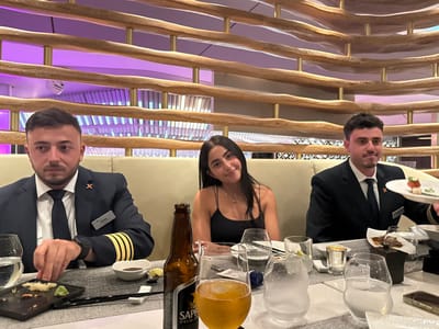 Three individuals sit at a table in a warmly lit restaurant. The two men flanking a woman are wearing dark uniforms with gold detailing. Sushi and beer are visible on the table, along with various dining utensils. A decorative wooden structure serves as a backdrop, adding depth to the setting. The woman is looking at the camera with a smile. The ambiance suggests a formal yet relaxed dining experience.