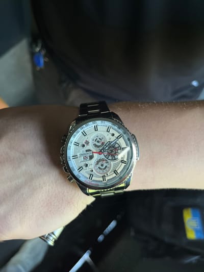 A close-up shot shows a person's wrist wearing a silver and white chronograph watch with a metallic band. The watch face displays multiple sub-dials, Roman numerals, and tachymeter markings. The background is blurred, but a dark fabric garment is visible, along with a hint of a blue keychain. The scene appears to be indoors, possibly in the city of Sviatopetrivske, Ukraine, suggested by a partial view of a blue and yellow Ukrainian symbol in the lower right corner of the frame.