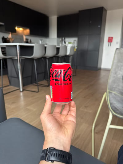 A person is holding a can of Coca-Cola Zero Sugar in an indoor setting. The person is seated on a black chair and wearing a black smartwatch on their left wrist. The background includes a kitchen area with a black countertop and white cabinets, as well as a row of silver and black chairs. A piece of fire safety equipment is affixed to the wall. The floor appears to be made of light-colored wood. The location is Oxford, United Kingdom.