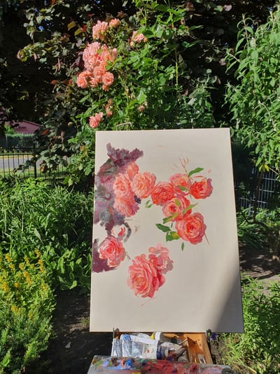This media file is a photograph of an oil painting of a bouquet of roses on a canvas. The painting is placed outdoors on a sunny day, surrounded by lush green foliage and a blooming rose bush. The roses in the painting are depicted in various shades of pink and red, with loose brushstrokes that give them a vibrant and impressionistic feel. Some of the roses are fully bloomed, while others are still in bud. The canvas is light beige and appears to be propped up on an easel or a similar support. To the right of the canvas, a wooden box filled with art supplies, including tubes of paint and brushes, is visible. The scene is set in a garden, with the natural beauty of the flowers and greenery serving as both a backdrop and inspiration for the artwork. The sunlight casts shadows on the canvas and the surrounding area, indicating it is likely midday. The overall mood is peaceful and creative, capturing a moment of artistic expression in a natural setting. There is no visible text in the image.