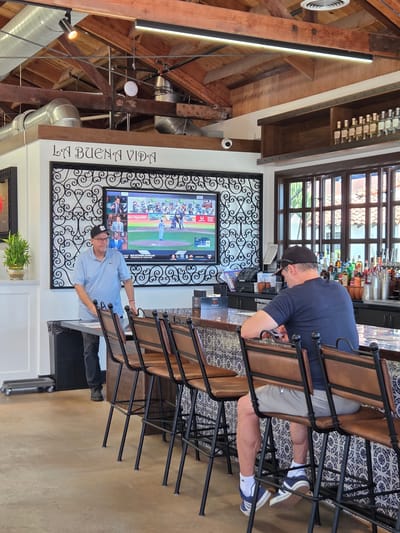 In San Clemente, United States of America, a restaurant interior features a bar area with people. A man in a light blue polo shirt and a baseball cap stands behind a counter, looking towards a large television screen displaying a baseball game. To his right, another man sits at the bar, facing away from the camera, wearing a dark blue t-shirt and light-colored shorts. The bar is lined with several empty brown leather bar stools. Behind the bar, shelves are stocked with various bottles of alcohol, and a large window with multiple panes is visible. The restaurant has an exposed wooden ceiling with industrial-style lighting and ductwork. The walls are decorated with the phrase "LA BUENA VIDA" in ornate lettering and a decorative metal screen.