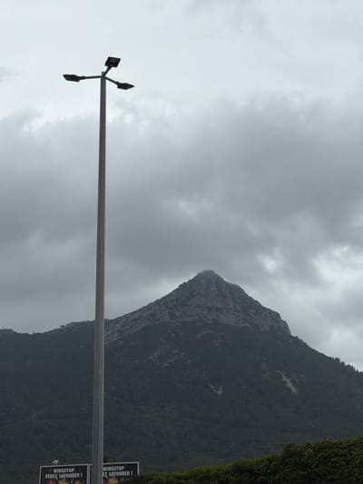 The image shows a vertical view of a cloudy sky over a mountain in La Garde, France. A tall light pole is on the left, and a sign advertising "Wingstop, Venez Savourer!" is on the right. The overall scene is overcast.