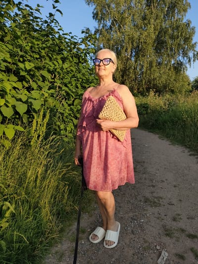 An elderly woman with blonde hair and glasses stands on a dirt path next to a dense green bush. She is wearing a pink, floral print dress and white slides. In her right hand, she holds a woven straw clutch. A black cane is visible by her left side. Tall trees are in the background, and the sky is clear and blue, suggesting it is daytime. The scene appears to be outdoors in a natural setting, possibly a park or rural area, with dappled sunlight suggesting late afternoon or early morning. The woman has a neutral expression. There is no visible text in the image.