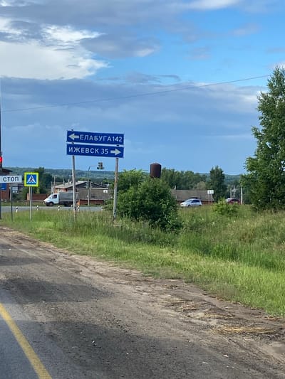The image shows a road scene in Minderevo, Russia, with directional signs indicating routes to Yelabuga (142 km) and Izhevsk (35 km). The sky is partly cloudy, with a mix of blue sky and white clouds. On the left side of the road, there is a red traffic light and a "STOP" sign, along with a pedestrian crossing sign. A white truck is visible driving on the road. In the background, there are several buildings, trees, and a water tower. The foreground shows a dirt road with some greenery. The overall scene depicts a rural road with traffic and signage.