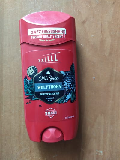 The image shows a red Old Spice Wolfthorn deodorant stick standing upright on a wooden surface. The deodorant stick features a prominent label with a wolf illustration, the Old Spice logo, and text indicating "WOLFHORN", "SCENT OF WILD CITRUS", and "DEODORANT STICK". Above this, it says "XXLLLL" and "24/7 FRESSSHHH PERFUME QUALITY SCENT". There is also a "3X ACTIVE DEFENCE" badge. The background is a textured wooden surface. The image is taken from a slightly elevated angle, looking down at the deodorant stick. There are no people or animals visible. There is no information available to indicate this image is located in the city of Turka, Ukraine.