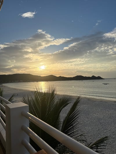 This is a wide shot of a beach in Hòa Hiệp, Vietnam, at sunset. The sun is low in the sky, casting a warm glow over the scene and reflecting on the water. In the foreground, a white railing and palm fronds partially obscure the view of the beach and ocean. The sand on the beach is light-colored and appears undisturbed, with a few scattered figures visible in the distance. The ocean is calm, with small waves lapping at the shore. Several boats are anchored in the water. In the background, rolling hills covered in vegetation form a silhouette against the sky. The sky is a mix of blue and white clouds, with the sun peeking through them. The overall atmosphere is tranquil and serene.