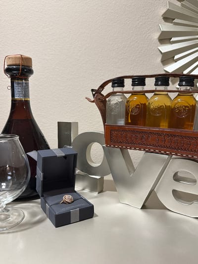In Riverview, United States of America, on a neutral-colored shelf, a scene is set with a large bottle of amber liquid, a brandy snifter, an open ring box displaying a diamond ring, and a leather-bound carrier holding four miniature bottles of alcohol. A decorative metal "LOVE" sculpture stands behind these items, and a starburst-shaped decorative piece is visible on the wall in the background. The lighting appears to be artificial, casting shadows on the shelf.