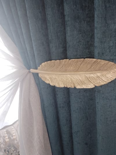 The image shows a close-up of teal-colored curtains with a gold feather-shaped curtain tieback. A sheer white curtain is visible on the left, with pearl embellishments on a darker patterned fabric behind it. The scene appears to be indoors, possibly in a residential setting. There are no people or signs of activity in the image. The location in the city of Октябрь, Kyrgyzstan, is not ascertainable from the image.