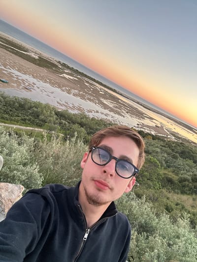 The image is a selfie taken outdoors, possibly in Vila Real de Santo António, Portugal. A young man with short brown hair, a light beard, and wearing black-rimmed glasses is the central figure. He is wearing a dark jacket that is partially zipped up. He appears to be standing on a slightly elevated position, possibly a rocky outcrop, surrounded by low-lying green vegetation. In the background, there is a view of a flat, sandy landscape interspersed with bodies of water, likely a coastal area or river delta. The sky in the distance shows a gradient of colors, suggesting either sunrise or sunset. The overall composition is oriented at an angle, giving the impression of a dynamic, informal snapshot.