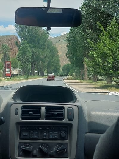 The image, taken from inside a vehicle, presents a forward view of a two-lane asphalt road leading into the distance, purportedly in Getap, Armenia. The car's dashboard, with grey and beige panels, occupies the lower third of the frame, showing air vents, a car stereo with Japanese text, and climate control knobs. A rearview mirror is partially visible at the top center.
On the left side of the road, a red gas station sign is clearly visible, displaying "ԳԵՏԱՊ" (Getap) and "LPG" with associated prices. A white truck is parked behind the sign on the shoulder. Both sides of the road are lined with a dense array of tall, leafy green trees. Beyond the trees on the left, a distinct reddish-brown rocky hillside rises under a bright sky with scattered white clouds. Further down the road, a red car is observed driving away in the right lane. The right side of the road also features a grassy verge and distant green hills. The environment appears rural or semi-rural, characterized by mountainous terrain and lush vegetation.
