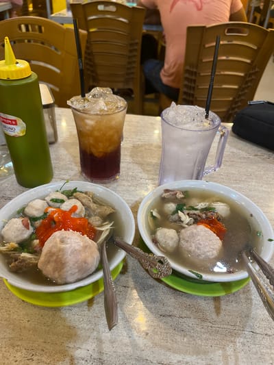 The image captures a close-up view of two bowls of bakso, a traditional Indonesian meatball soup, placed on a textured table at what appears to be a casual dining establishment. The bowls are white and oval-shaped, each filled with a clear broth containing large meatballs, noodles, and some leafy greens. One bowl is topped with a dollop of bright red chili sauce. A green squeeze bottle, likely containing sauce, stands to the left of the bowls, with a yellow cap and a label that reads "LION STAR BRUNO SAUCE KEEPER." Two tall glasses, one filled with ice and a dark beverage, and the other with ice and a clear liquid, are also visible. The background shows blurred wooden chairs and the torso of a person wearing a pink t-shirt, suggesting an indoor seating area. The overall lighting is warm, indicating ambient restaurant lighting. The scene is set in Kampongbaro, Indonesia.