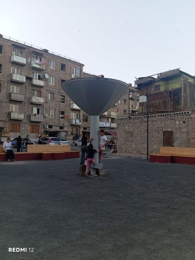 In the city of Vedi, Armenia, an outdoor scene unfolds with a backdrop of residential buildings and a distressed wooden structure. In the foreground, a modern, umbrella-like sculpture dominates the space, with a few children interacting with it. One child, a girl in pink pants and a dark top, is climbing up the central pole of the sculpture, while another child, a boy in a light-colored shirt, is also on the sculpture. To the left, a woman with short dark hair sits on a red-bordered bench, holding a light-colored object, possibly a ball. Several other individuals are visible, some standing near a silver car parked in front of the buildings. The ground is covered with dark, gravelly material, and wooden benches with red borders are placed around the area. The buildings in the background display a mix of architectural styles, with one appearing to be an older, multi-story apartment building with balconies and laundry hanging out, while the other is a more rudimentary, weathered wooden structure. The sky is overcast.