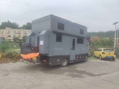 The image shows a large, grey recreational vehicle (RV) parked on a gravel and dirt lot. The RV appears to be custom-built and has a boxy design with several windows and compartments. A smaller yellow car is parked to the right of the RV, and a motorcycle covered with a black and orange tarp is attached to the rear of the RV. The background consists of trees and buildings, suggesting an urban or suburban setting. The sky is overcast. The license plate on the yellow car reads "川G C167K". The license plate on the RV reads "川E 73319". The RV has a logo on its side that appears to be a crest. In the distance, there is a solar-powered street light. This scene is located in Jianyang City, China.
