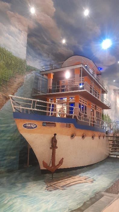 The image displays an indoor installation in Glantengan, Indonesia, meticulously designed to simulate an outdoor maritime setting. The focal point is a large, multi-deck boat structure, appearing approximately three stories in height. The boat features a cream-colored hull, a distinct blue stripe along its upper section, orange support pillars, and silver handrails on each level. A reddish-brown anchor is visibly suspended from the bow, which also displays the name "MUARAH" in stylized script. Several circular portholes are present along the boat's side. The interior, visible through windows, is illuminated with a blue glow, and an ornate, golden-colored object is discernible on the second deck. The floor is painted with light blue and green hues to represent rippling water, with a depiction of a wooden raft or debris floating near the boat's bow. The walls and ceiling are adorned with murals portraying a sky filled with clouds and a natural landscape, including lush green vegetation resembling palm trees along a riverbank on the left side of the scene. Multiple bright, artificial light sources are visible, illuminating the installation. No individuals are present in the image.