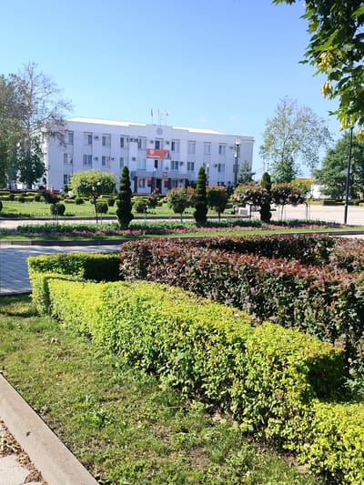 The image depicts a bright, sunny day with a clear blue sky above a two-story, white, modern building. The building has numerous windows and air conditioning units, and a large orange banner with Russian text, possibly an advertisement or a slogan, is visible on its facade. Three flags, appearing to be Russian, fly from poles on the roof of the building.
In front of the building, there is a manicured garden with well-kept bushes and trees. Some trees are shaped into topiaries, including conical and spherical forms, with some adorned with pink flowers. Flowerbeds with small pink and purple flowers are also visible. A sidewalk or paved area is present in the foreground, bordered by neatly trimmed hedges. The overall scene suggests a public space, likely in a city, with a focus on civic order and landscaping.
Based on the context, this image is of the administration building in Labinsk, Russia.