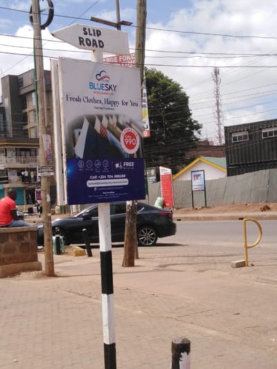 The image displays a street scene within Kikuyu Township ward, Kikuyu, Kenya, specifically near the location indicated on a billboard as "Matasia Mall, Kikuyu Town (Rail Road)." A prominent vertical billboard for "BLUESKY DRYCLEANERS LTD" occupies the central foreground, advertising "Fresh Clothes, Happy for You" and a promotion for "10 ITEMS FOR KShs 990" with "FREE Pick & Drop." The billboard includes contact details: phone number +254 704 588588 and email samuel@blueskydrycleaners.co.kz.

Above the billboard, a white directional sign with a black arrow points left, reading "SLIP ROAD." To the left, an adult male in a red short-sleeved shirt and blue jeans is seated on a low stone wall. A dark-colored sedan is parked on the side of the road, facing right. Multiple utility poles and overhead wires crisscross the frame. On the far right sidewalk, a yellow P-shaped metal structure, likely a parking barrier, is anchored to the ground. In the middle ground on the right, a corrugated metal fence borders a property with a building featuring a light yellow-green roof and white walls. A tall cellular tower is visible in the distant background against a partly cloudy sky. Multi-story buildings are present in the background on the left, with several indistinct figures visible on a balcony.