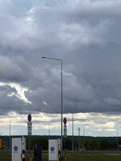 The image shows an outdoor scene in Gil'deevo, Russia, under a dramatic sky with contrasting light and dark clouds. Along a roadside, there are street lamps and regulatory road signs, including a "no stopping" symbol. Two electric vehicle charging stations are visible, branded with "Сетевая Компания." A green strip of vegetation, possibly a forest edge, is present in the background.