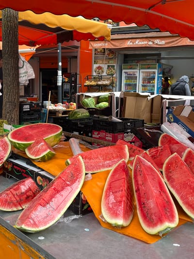 At a street-side fruit stand in Villeparisis, France, a variety of watermelons are on display. Several large, halved watermelons are prominently featured, with some cut into wedges and wrapped in plastic. Surrounding these, whole watermelons are visible, some in black plastic crates. The stand is situated outdoors, beneath a bright orange and yellow awning. In the background, a storefront with refrigerators stocked with beverages is visible, along with a sign that reads "La Maison du Fruits." A person wearing a grey hooded jacket is partially visible in the background, standing near the storefront. The scene appears to be a typical market setting with fresh produce being sold.