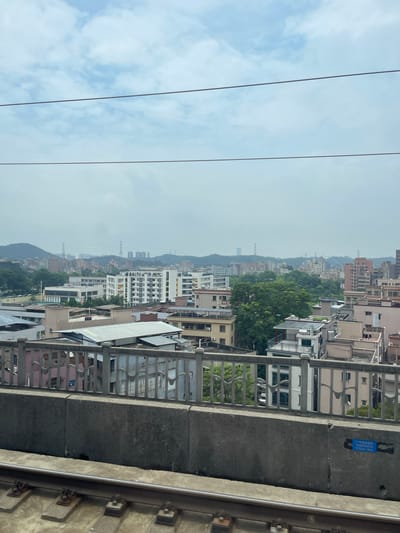 This is an eye-level, medium shot of a city skyline in Dongguan, China, taken from a moving train. In the foreground, train tracks run horizontally across the bottom of the frame. A concrete barrier with a metal railing separates the tracks from the city beyond. The city is densely populated with a mix of multi-story residential buildings, some with balconies and solar panels, and a few larger apartment complexes. Green trees are interspersed throughout the urban landscape, and in the distance, hills rise under a hazy, overcast sky. The sky itself is a pale blue with scattered white clouds and appears to be divided by two power lines that stretch across the upper portion of the image. There are no people visible in the image.