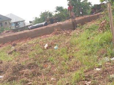 The image shows a construction site in Achara Ndiagu Omegio, Nigeria. A partially constructed building foundation is visible, with reinforcing bars extending upwards. A large, black, cylindrical object rests on the foundation. Several people are present at the site. One man in a red shirt is standing on a ladder, and others are nearby. Bricks are stacked near the foundation. In the foreground, the ground is covered in a mix of dirt, dry grass, and green vegetation. A white chicken and a blue plastic object are visible in the field. A finished building is in the background, with gray roofing. The sky appears overcast.