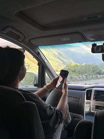 The image shows a person sitting in the passenger seat of a car, looking at their phone. The car is traveling on a road that winds through a mountainous landscape. The person is wearing a grey t-shirt and grey pants, and has a pink watch on their wrist. The landscape outside the car is characterized by lush green mountains with some trees. The sky is bright and appears to be late afternoon or early evening, with the sun casting a warm glow on the mountains. The dashboard of the car is visible, with a screen and various buttons. The interior of the car suggests it is a modern vehicle. The specific location in the city of Chmi, Russia is not explicitly identifiable from the provided image.