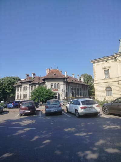 The image depicts a street scene in Trușești, Romania, featuring two prominent buildings and several parked cars. The building on the left is a multi-story structure with a red tiled roof, ornate chimneys, and numerous arched windows. A Romanian flag is visible on a flagpole in front of this building. To its right, a second, lighter-colored building with a similar architectural style and a mansard roof is visible, adorned with decorative elements. In the foreground, a parking lot is occupied by several vehicles: a dark blue SUV, a burgundy hatchback, a silver hatchback, a white station wagon, and a dark gray sedan. The ground is asphalt, with white parking space markers visible. The sky above is clear and blue, suggesting a sunny day. The overall scene suggests a quiet urban or suburban environment.