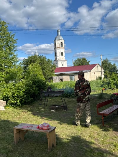 This outdoor image, captured on a sunny day in what appears to be a rural or village setting, depicts a man tending to a grill. The man is positioned slightly right of center, seen from behind and to his left, wearing camouflage trousers and a matching long-sleeved shirt with a cap. Smoke gently rises from a black metal grill or brazier in front of him, suggesting he is cooking.
The ground is covered in lush green grass, with patches of darker, possibly worn earth. To the man's right, a rustic wooden bench or table holds a clear plastic bag filled with red vegetables, possibly tomatoes or peppers, alongside a red spatula. Another empty wooden bench with red-painted legs is visible further back and to the right. Green trees and dense foliage form a natural backdrop across the scene.
In the background, a distinctive white bell tower of a church with a silver dome rises prominently on the left side of the frame. Another smaller, silver-domed structure is partially visible behind the trees further left. A building with a red roof and light-colored walls is situated behind the man, with more greenery and other structures in the distance. The sky is bright blue with scattered white cumulus clouds, and power lines stretch across the upper portion of the image. The overall atmosphere suggests a relaxed, summer day activity in a traditional Russian village setting, consistent with the provided location of Луптюг, Russia. No visible text is present.