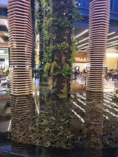 The image depicts the interior of a shopping mall in Araçariguama, Brazil. In the foreground, a shallow body of water with visible pebbles and reflections of lights and structures occupies the lower portion of the frame. Two tall, octagonal wooden pillars with a layered design flank a central vertical garden structure. The garden is composed of dark fabric pockets containing a variety of green plants, some of which are trailing downwards. In the mid-ground and background, shoppers are visible seated at tables in a dining area with a sign that reads "GRILL & SAFARI." The environment appears well-lit with artificial lighting, and the overall atmosphere is that of a modern, indoor public space incorporating natural elements.