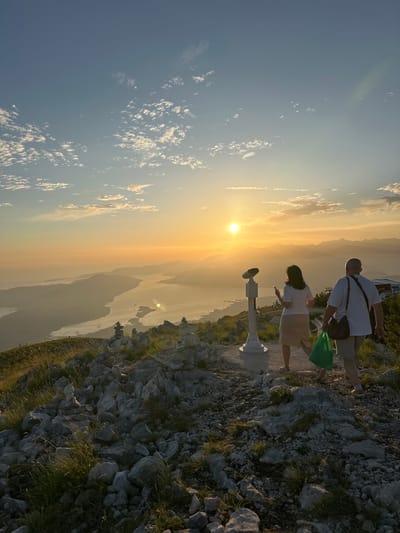 This is a photograph taken at sunset from a high vantage point, likely the peak of a mountain overlooking the Bay of Kotor, near the city of Mirac, Montenegro. The sky is a gradient of soft oranges, yellows, and blues, with scattered, wispy clouds. The sun, a bright orb, is low on the horizon, casting a warm glow over the scene.

In the foreground, the terrain is rocky and covered with sparse, dry grass. Several cairns, stacks of stones, are visible on the ridge, suggesting this is a popular viewpoint. Two people are walking away from the camera, heading towards the viewpoint. The person on the left appears to be a woman wearing a light-colored skirt and top, holding a smartphone. The person on the right is a man in a white t-shirt and light-colored shorts, carrying a brown bag over his shoulder and a green plastic bag in his hand.

Further down the slope, a coin-operated telescope is mounted on a pedestal, indicating a tourist attraction. Beyond the immediate ridge, the expansive Bay of Kotor stretches out, dotted with islands and surrounded by misty, silhouetted mountains. The water reflects the sunset light. In the distance, faint structures and possibly more cairns can be observed on other elevated points. The overall atmosphere is serene and picturesque, capturing a moment of natural beauty and human presence at a scenic overlook.
