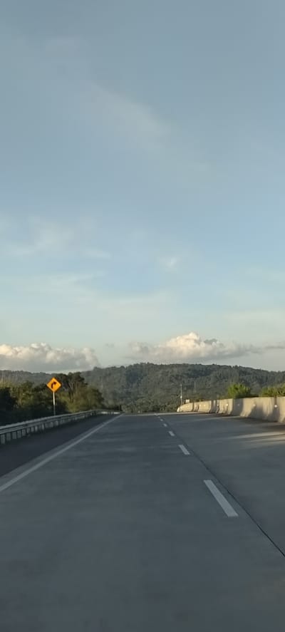 The image shows an empty highway with a gentle curve to the right, surrounded by lush green hills under a pale blue sky with scattered clouds. On the left side of the road, a yellow diamond-shaped warning sign with a black arrow indicating a curve is visible. The road surface is asphalt, with clear white lane markings. Concrete barriers line the right side of the highway. The scene suggests a rural or mountainous area. There are no people or vehicles visible in the image, indicating a quiet and serene environment. The lighting suggests it is either early morning or late afternoon, with soft sunlight casting a gentle glow on the landscape. The location in Lonbaroh, Indonesia, would imply a tropical climate, consistent with the abundant greenery.