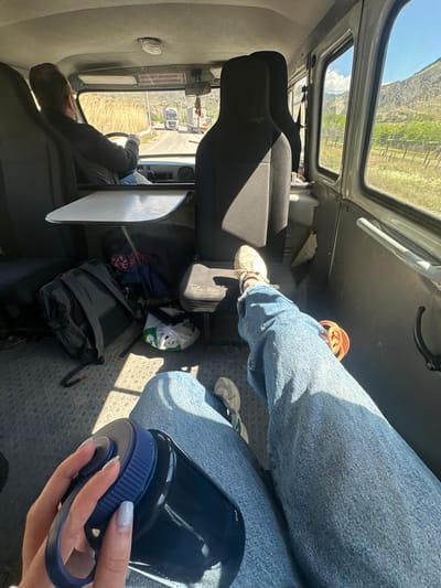 The image shows the interior of a vehicle, likely a van or bus, viewed from a passenger's perspective during bright daylight. Strong sunlight streams into the cabin, casting distinct shadows.
In the foreground, a person's legs are visible, clad in blue jeans and white sneakers. Their hand, adorned with light-colored nail polish, holds a dark blue water bottle with a matching blue lid and strap. Another passenger is seated further back, their legs extended, also wearing blue jeans and white sneakers; orange sandals are visible near their feet. On the floor between the seats, a black backpack and a white plastic bag with green lettering are present. A small, light-colored fold-down table is positioned to the left.
The driver is partially seen from behind in the front, wearing a dark shirt and holding the steering wheel. Through the front windshield, a road with dry grass or fields can be seen, and other vehicles, possibly trucks, are visible in the distance. The vehicle's roof features skylight windows, revealing a clear blue sky with some clouds and a distant, mountainous landscape with arid terrain and green patches, consistent with the Meghri region of Armenia.
Above the driver's head, a white sign displays Cyrillic text: "ЧИСЛО МЕСТ ДЛЯ СИДЕНИЯ" (Number of seats for sitting) and "ОБЩЕЕ ЧИСЛО ПАССАЖИРОВ" (Total number of passengers), each followed by the handwritten number "8". The interior floor is covered with black rubber matting featuring a circular studded pattern. The scene depicts a journey through a scenic, possibly mountainous area.