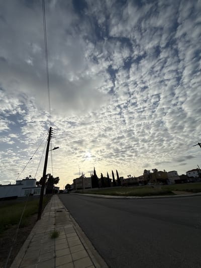 The image depicts a street scene in Paralimni, Cyprus. The view is taken from street level, looking towards a residential area. The sky is filled with altocumulus clouds, with the sun positioned behind them, creating a diffused light. A wooden utility pole with attached electrical wires is visible on the left side of the frame. The street surface is asphalt, with a concrete sidewalk on the right. Buildings, primarily residential, line the street. Trees are interspersed between the buildings. The lighting suggests either early morning or late afternoon. There are no people visible in the scene.