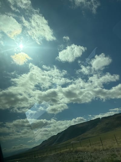 This is an outdoor, eye-level, full-shot photograph taken from inside a vehicle, showing a sun-drenched sky filled with fluffy white and grey clouds. The sun is visible in the upper left quadrant, casting a bright glare. A slanted, barren hillside with patches of dry grass and a few scattered green areas slopes down from the upper right to the lower left of the frame. A dark, wire fence runs along the base of the hill. Reflections of clouds and the sky are visible on what appears to be a car window in the foreground, obscuring a portion of the hillside and sky. The scene is captured from a moving vehicle, as indicated by the reflections and motion blur. The location is identified as Zhansugurov, Kazakhstan. There are no people or animals present in the image.