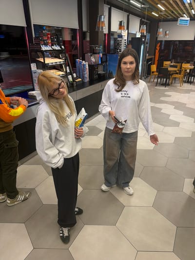 Two women are standing on a gray tiled floor inside a commercial building. The brunette wears a white, long-sleeved shirt, blue jeans, and white sneakers; she holds a bag of chips and a mobile phone. The blonde wears a light gray sweatshirt and black pants, and holds a bag of chips. Another person, wearing an orange and brown shirt and green pants, is visible in the lower part of the frame. The location appears to be a store or cafe, as evidenced by the items displayed behind the women. The hexagonal floor tiles suggest a modern interior. The image has an unusual orientation, with the subjects and setting rotated approximately 90 degrees. The city of Stara Pazova Municipality, Serbia is not discernible from the image.