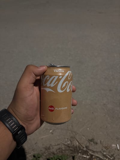 A person's hand, wearing a black smartwatch, holds a can of Coca-Cola Vanilla Flavors. The can is tan colored with white lettering, including "Vanilla" at the top and the Coca-Cola logo. The can appears to be cold, with condensation visible. The background is a blurred, gray, asphalt-like surface, with some dry grass and debris in the lower right corner. The lighting suggests it is either dusk or night.