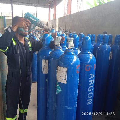 An indoor industrial facility in the city of Pakuro, Nigeria, depicts a dark-skinned male worker, positioned left-of-center, wearing a dark blue jumpsuit with reflective yellow-green stripes and a dark face mask. He is holding a teal/turquoise heat gun with both hands, directing its nozzle toward the upper section of a blue gas cylinder. The foreground and mid-ground are densely populated with numerous upright blue gas cylinders, many featuring white labels; one prominent cylinder has "ARGON" stenciled vertically in white with red outlining. The background consists of light-colored concrete block walls and an overhead metal truss structure, indicating an industrial roof. A small section of green foliage is visible in the upper right background, suggesting an exterior view. The scene is well-lit. A digital timestamp "2025/12/9 11:28" is overlaid in the lower right corner.