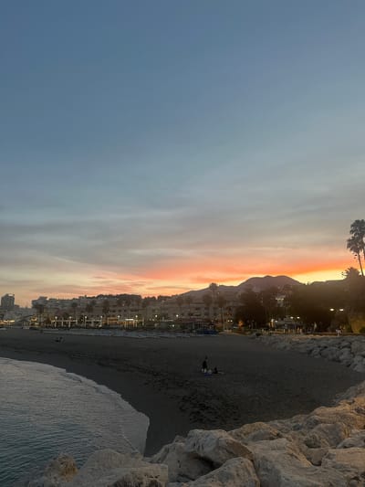 The image depicts a wide-angle view of a beach in Torremolinos, Spain, at dusk. The sky displays a gradient of colors, transitioning from a pale blue at the top to vibrant orange and pink hues near the horizon, indicating the setting sun. In the background, a coastal town stretches along the shoreline, characterized by multi-story buildings, palm trees, and streetlights that begin to illuminate. The beach itself is composed of dark sand and is lined with rows of empty lounge chairs and umbrellas, suggesting it was recently occupied or is preparing for the next day. Several large rocks form a breakwater in the foreground, with the gentle waves of the ocean lapping against them. A few scattered individuals are visible on the beach, one pair seated on a blanket, and another person standing. The overall atmosphere is serene and captures the tranquil beauty of the Spanish coast as day turns into night.