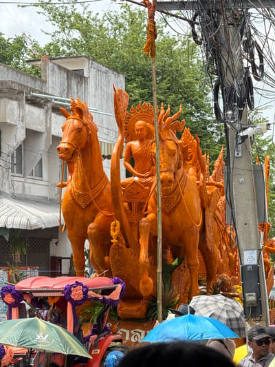 This is a photograph taken in the city of Prakhon Chai District, Thailand, depicting a parade float adorned with intricate orange wax sculptures. The float is pulled by a red tractor, decorated with purple and orange fabric flowers and streamers. In the foreground, people are visible, some holding umbrellas, including a green one on the left and a blue one in the center. The main subject of the float is a large, ornate structure featuring two horses and a seated figure, possibly a Buddhist monk, in elaborate orange wax carvings. The backdrop consists of buildings and lush green trees, with a prominent utility pole laden with numerous black electrical wires on the right side of the image. The sky appears overcast.