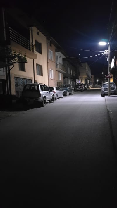The image depicts a dimly lit street at night in what appears to be a residential area of Breznitsa, Bulgaria. Several cars are parked along the side of the road, next to the buildings. A street light illuminates a portion of the street and the facades of the buildings. The road surface is dark, indicating that it's likely paved with asphalt. In the distance, another street light and a road sign are visible. The sky is dark.
