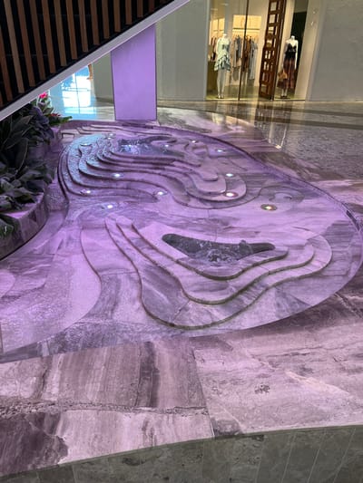 The image shows a water feature in the Indooroopilly Shopping Centre in Brisbane, Australia. The water feature is made of grey stone and features a series of cascading pools, creating a flowing effect. The water in the pools is illuminated by purple LED lights. There are several recessed lights surrounding the water feature. There is a small patch of green foliage at the bottom left of the image. The background of the image is out of focus, but it appears to be a shop front. The shop front is illuminated by warm-toned lights.  The scene is deserted and appears to have been captured on a quiet day. The image has a purple hue and a smooth texture.