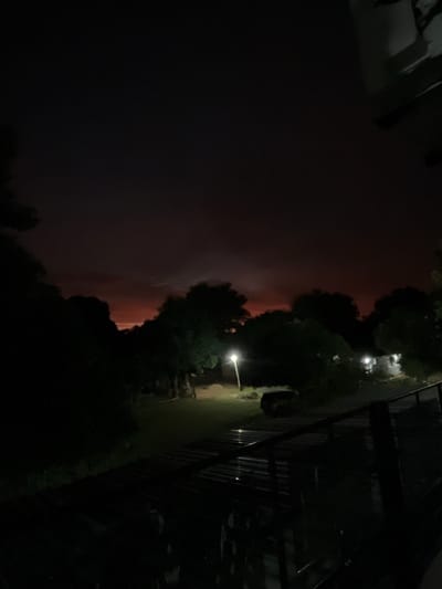 This is a nighttime, outdoor scene in Luque, Paraguay. The image appears to be taken from a slightly elevated position, possibly a balcony or window, looking out over a landscape. The most striking element is the sky, which is dominated by dark clouds illuminated from within by a deep reddish-orange glow. This suggests a dramatic atmospheric event, potentially a storm with lightning, or a distant fire reflecting off the clouds.

In the foreground and to the right, there is a dark silhouette of trees and foliage. A single, bright street lamp illuminates a small patch of grass and what looks like a fence or railing structure. Further back, amidst the trees, there are faint lights that suggest buildings or houses in the distance. The overall impression is one of a subdued, perhaps slightly ominous, nighttime scene, characterized by the contrast between the dark surroundings and the dramatic, colorful sky. The time of day is clearly night due to the artificial lighting and the darkness of the sky. There is no visible text in the image.