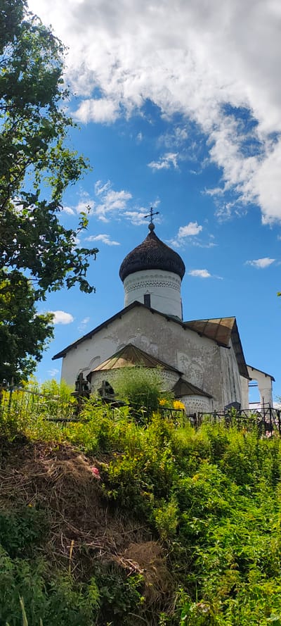 A low-angle, full-shot photograph captures the exterior of the Church of the Transfiguration in the village of Shchiglitsy, Russia, under a bright, partly cloudy sky. The church, constructed primarily of white stucco, features a dark, domed roof topped with a cross. Its facade displays signs of age and weathering. In the foreground, lush green vegetation and grass grow densely, interspersed with several weathered wooden crosses, suggesting the presence of a cemetery. A dark metal fence runs horizontally across the lower portion of the image, separating the viewer from the church and cemetery grounds. A large tree with dark green leaves frames the left side of the scene. The overall impression is one of a historical religious site nestled within a natural, overgrown landscape.