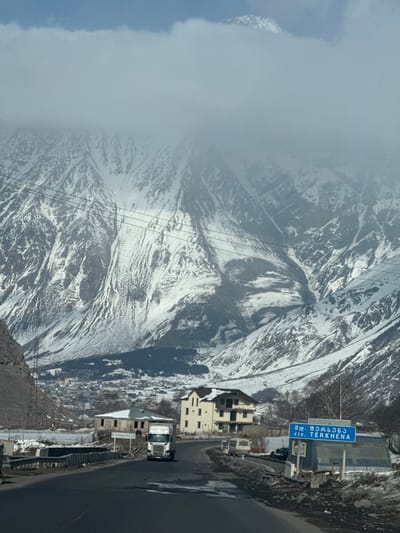 The image displays a winter mountainous landscape in the vicinity of Sioni, Georgia. A two-lane asphalt road, showing patches of residual snow or slush on its shoulders, curves through the scene. A white semi-truck with a trailer is positioned on the left lane, moving away from the viewer. To the right of the road, a blue rectangular road sign is visible, displaying Georgian script and the English text "riv. TERKHENA," indicating a local river or area. Several buildings, some with snow on their roofs, are situated alongside the road. A prominent multi-story, light-colored building stands on the right, while a stone structure, possibly incomplete, is on the left. In the mid-ground, a town or village is observed in a valley, backed by vast, snow-covered mountains that extend upwards. The upper portions of these mountains are partially obscured by low-hanging clouds. Overhead power lines cross horizontally through the mid-section of the image. The scene is devoid of discernible individuals.