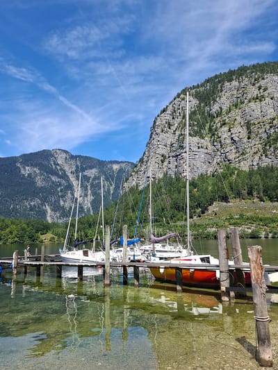 The image shows a picturesque scene at the harbor of Obertraun, Austria, on a sunny day with a bright blue sky adorned with wispy white clouds. The foreground features a wooden pier extending into a clear, shallow body of water, revealing the rocky lakebed beneath. Several sailboats are moored to the pier, their masts reaching towards the sky.

Most prominent are the sailboats, with white hulls and dark blue covers on their booms. One sailboat on the far right has a red, orange, and yellow hull. The boats are docked neatly, suggesting a tranquil and organized harbor.

The middle ground is dominated by a verdant green landscape, with trees lining the shore and ascending the steep slopes of a majestic mountain. The mountain face itself is characterized by dramatic rock formations, with streaks of lighter gray rock cutting through the darker, vegetated areas. The lush green trees contrast sharply with the rugged, rocky texture of the mountain.

In the background, more mountains, also covered in dense forest, rise higher into the sky. A small, solitary building with a red roof is visible nestled among the trees on the distant left shore, adding a touch of human habitation to the natural grandeur.

A single person, a male wearing a dark shirt and shorts, is standing on the pier near the edge, looking out at the water. His presence is minimal, emphasizing the vastness and serenity of the natural environment. The overall impression is one of peaceful coexistence between human activity and the awe-inspiring beauty of the Austrian Alps.