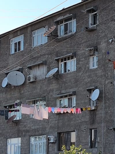 The image shows the exterior of a weathered, multi-story building, likely an apartment complex, in Şəki, Azerbaijan. The building's facade is rough and gray, showing signs of age and possibly incomplete renovations. A series of windows, some with intact frames and others appearing damaged or unfinished, punctuate the wall. Several satellite dishes are mounted on the exterior, indicating a common form of television reception in the area.

Draped across wires strung vertically down the building are various items of clothing, some brightly colored and others in pastel shades, suggesting laundry is being air-dried. These clothes add a touch of everyday life to the otherwise stark facade. A few small, possibly decorative items are also visible hanging on the wires, adding to the lived-in feel.

The angle of the photograph suggests it was taken from a low vantage point, looking upwards towards the building. The sky, visible in the upper left, is a clear blue, hinting at fair weather. The overall impression is one of a functional, perhaps somewhat impoverished, urban environment. There is no visible text in the image. The time of day appears to be daytime, with natural light illuminating the scene.